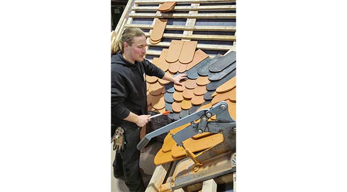 Ein Handwerker arbeitet an einem Dach und befestigt unterschiedlich geformte Dachziegel in verschiedenen Farben auf einer Holzkonstruktion.