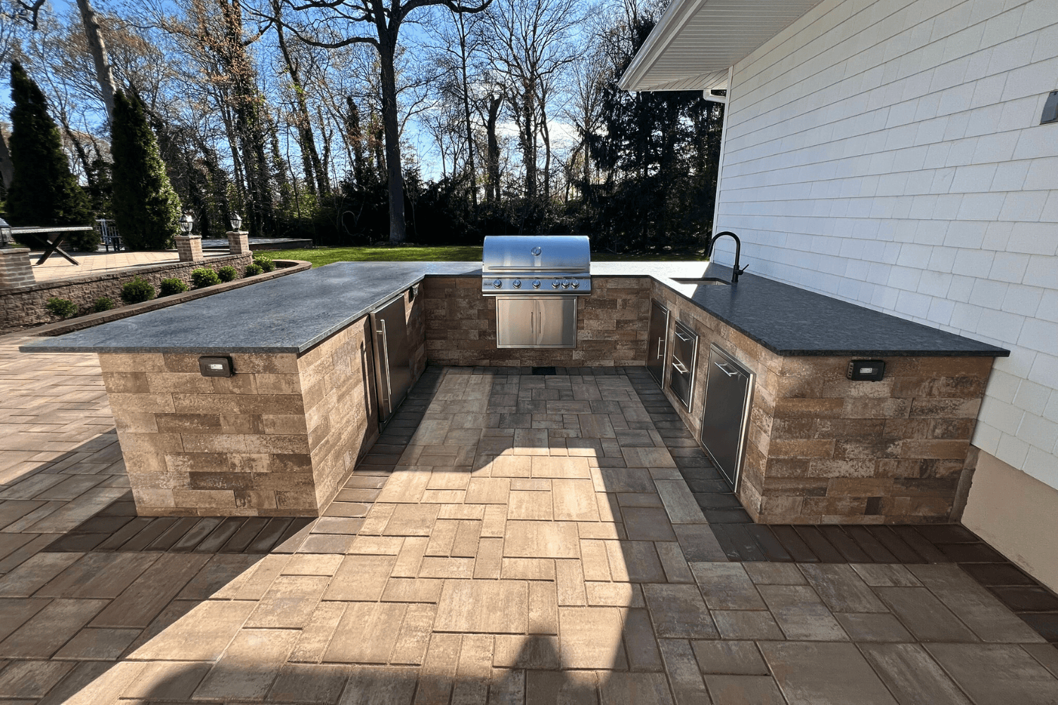 Outdoor Kitchens shine in this space with stone countertops, a built-in grill, stainless steel appliances, sink, and ample counter space on a paved patio. Trees and lawn create a lush backdrop.