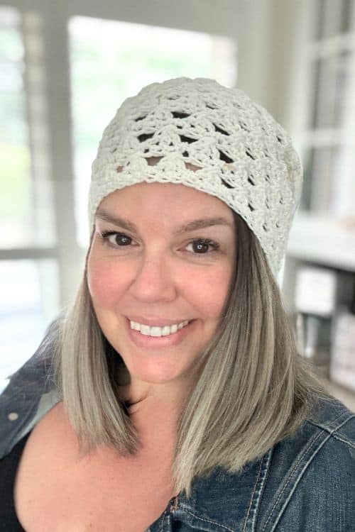 A woman with straight, shoulder-length hair smiles at the camera while wearing a simple granny stitch crochet beanie and a denim jacket, indoors with light streaming through windows in the background.