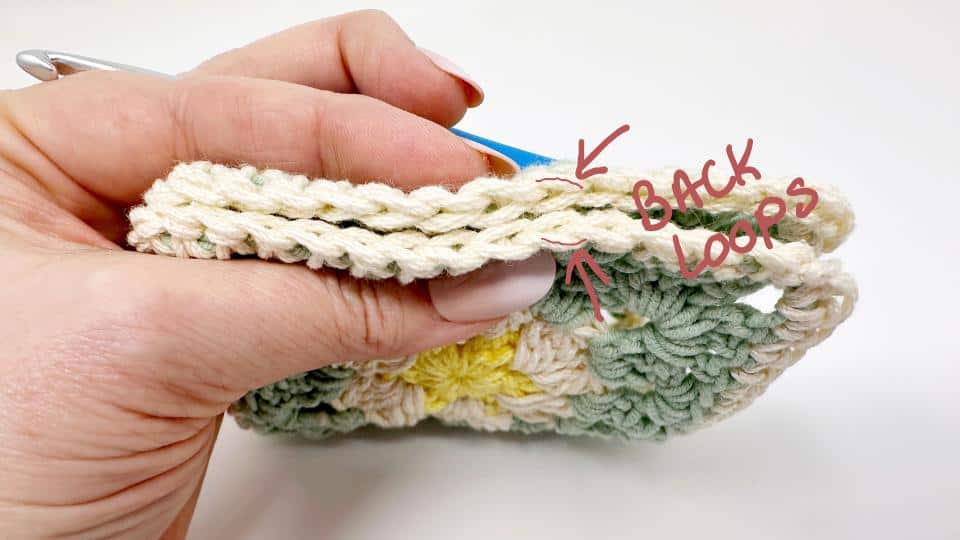 A hand holds a crochet project, showing a close-up of the stitches. An arrow highlights the BACK LOOPS of the top row—perfect for those working on granny square placemat free patterns and learning where to insert the hook.