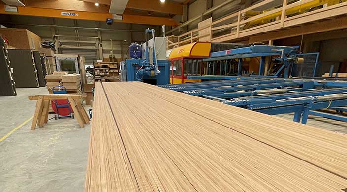 Holzplatten auf einem Förderband in einer Fabrikhalle mit Maschinen und Anlagen im Hintergrund.