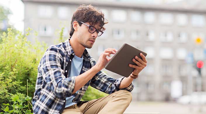 Ein junger Mann mit Brille und kariertem Hemd sitzt draußen und benutzt ein Tablet.