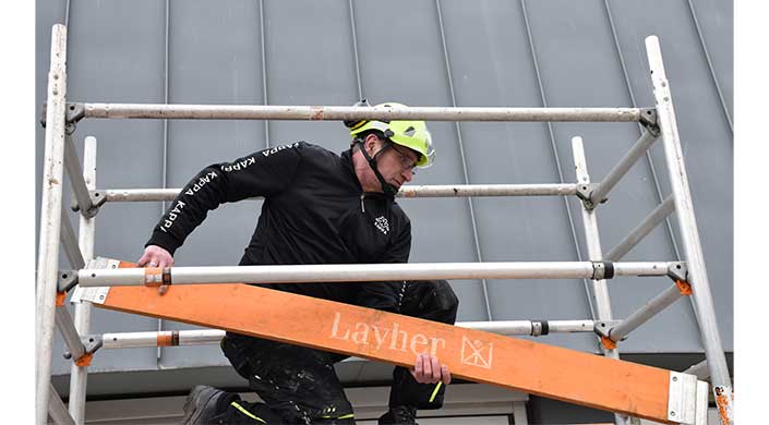 Ein Bauarbeiter mit Schutzhelm hält ein Layher-Brett und arbeitet auf einem Gerüst vor einem modernen Gebäude.