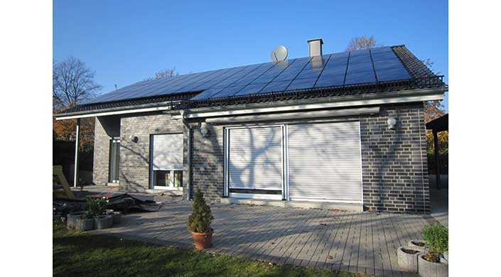 Modernes Haus mit Solarpanelen auf dem Dach, geschlossene Rollläden an den Fenstern, Satellitenschüssel und Schatten eines Baumes auf der Fassade.