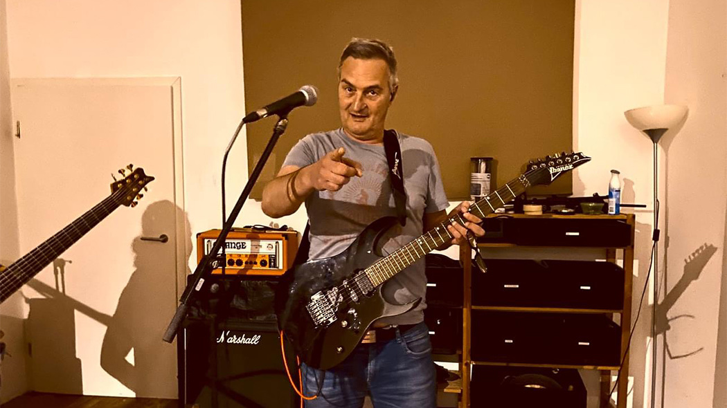 Ein Mann mit Gitarre zeigt in die Kamera und trägt ein graues T-Shirt.
