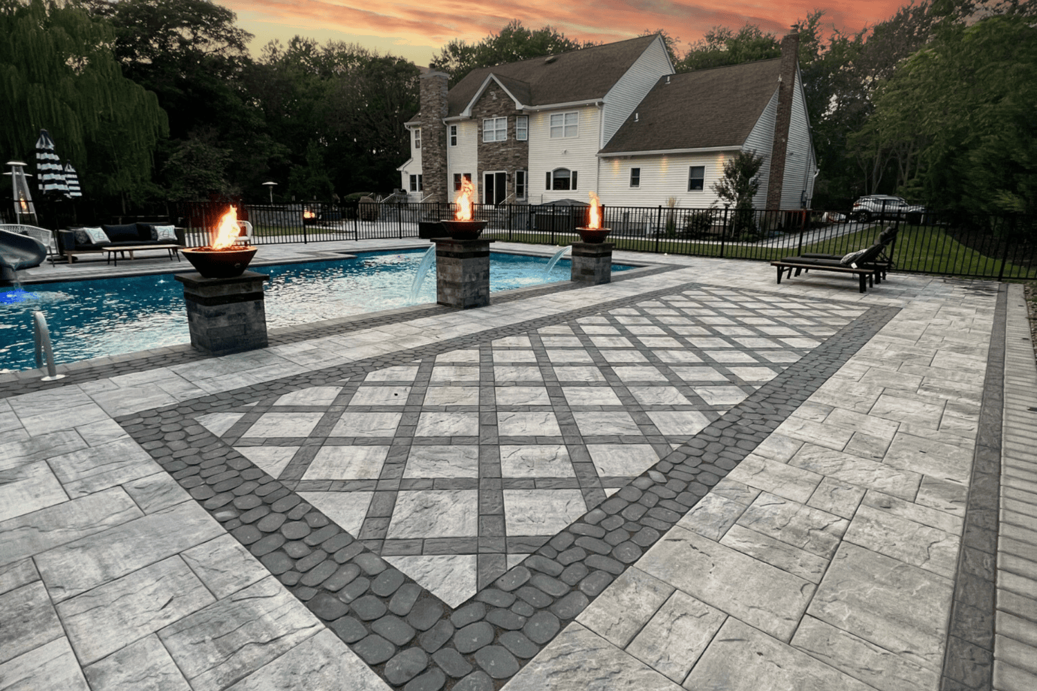 Backyard pool area with stone patio, geometric tile design, three fire bowls on pedestals, lounge chairs, and a spacious house in the background at sunset—perfect for relaxing evenings or entertaining on elegant patios.