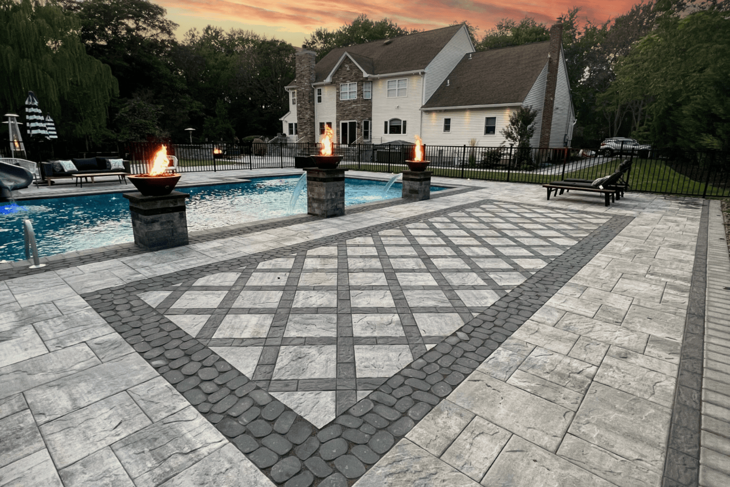 Spacious backyard patios feature geometric stone patterns, three fire bowls, a shimmering pool, lounge chairs, and a large house in the background illuminated by the warm glow of sunset.