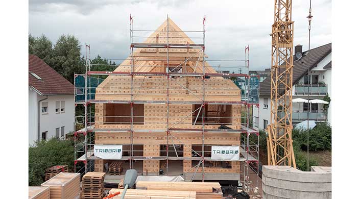 Ein Holzhaus im Bau mit Gerüst, sichtbaren Holzbalken und einer dreieckigen Dachform.