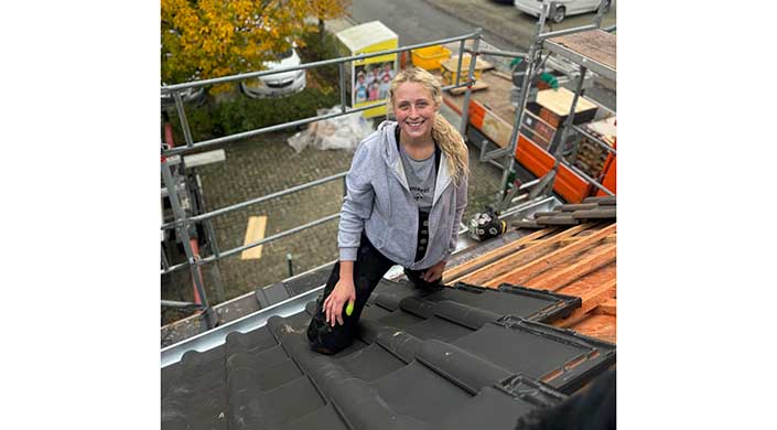 Eine Frau mit blonden Haaren und grauer Kapuzenjacke kniet lächelnd auf einem Dach, im Hintergrund Baugerüste und Baumaterialien.