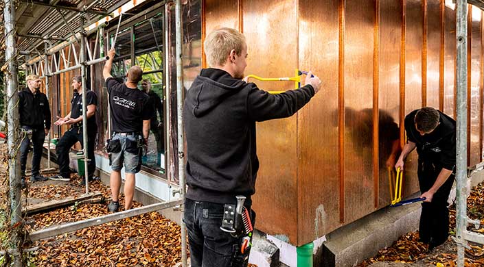 Ein Mann mit Werkzeug am Gürtel arbeitet an einer Holzwand und misst mit einem gelben Maßband.