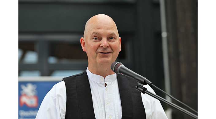Ein glatzköpfiger Mann im Anzug spricht draußen in ein Mikrofon und lächelt leicht.
