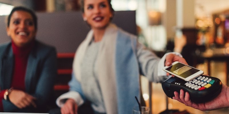 Two women are sitting in a cozy indoor space, smiling and conversing, while a hand extends a payment terminal. One of the women holds out a smartphone to complete a contactless payment, illustrating modern and seamless mobile payment technology in a social setting.
