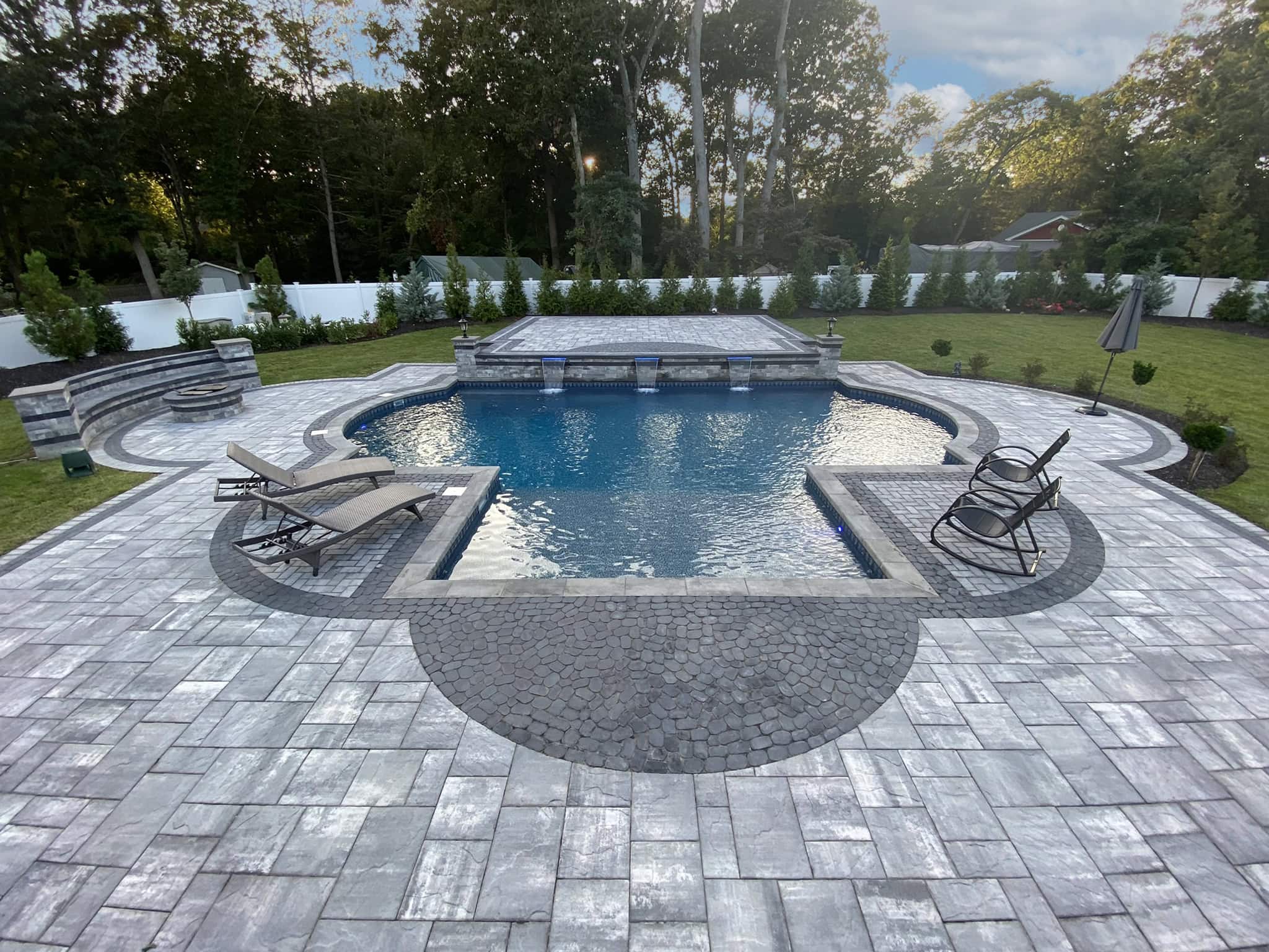 A backyard swimming pool with two lounge chairs, surrounded by stone pavers and greenery, with a hot tub at the far end and trees in the background.