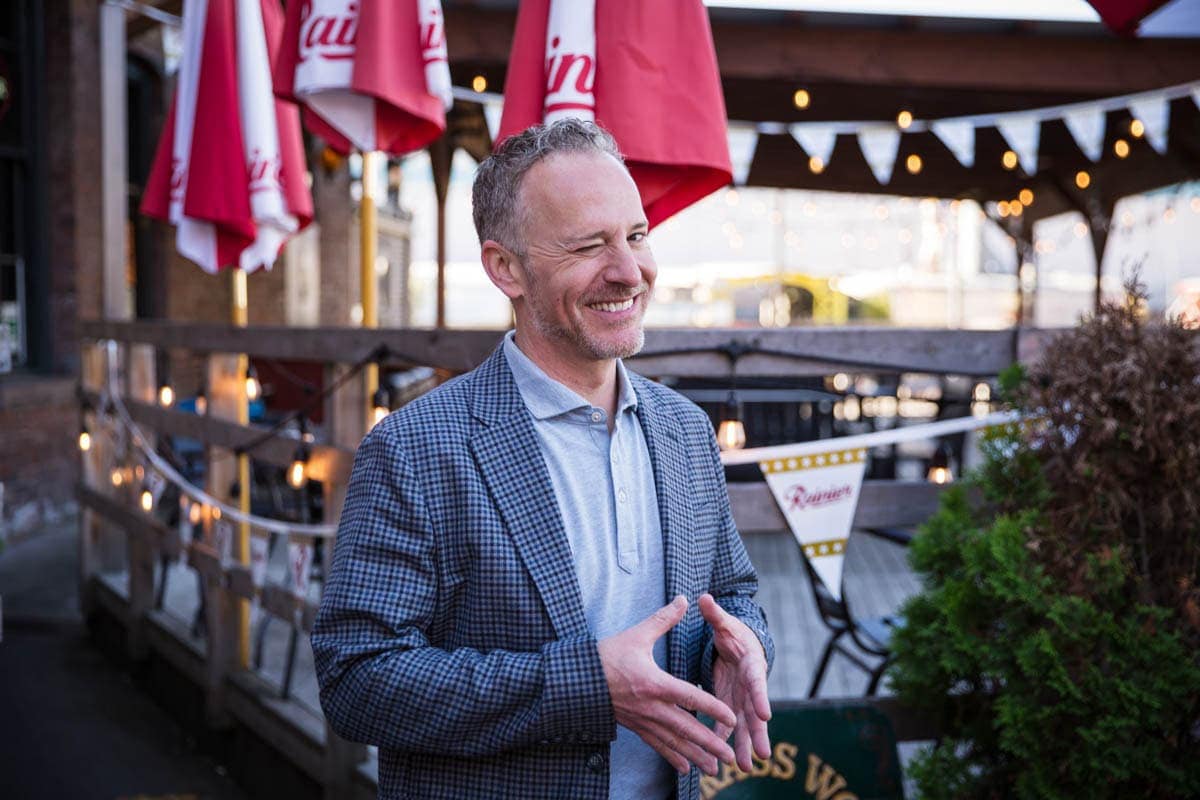 A man in a plaid blazer and light gray polo shirt stands outdoors near a patio with red umbrellas and string lights, smiling and gesturing with his hands.