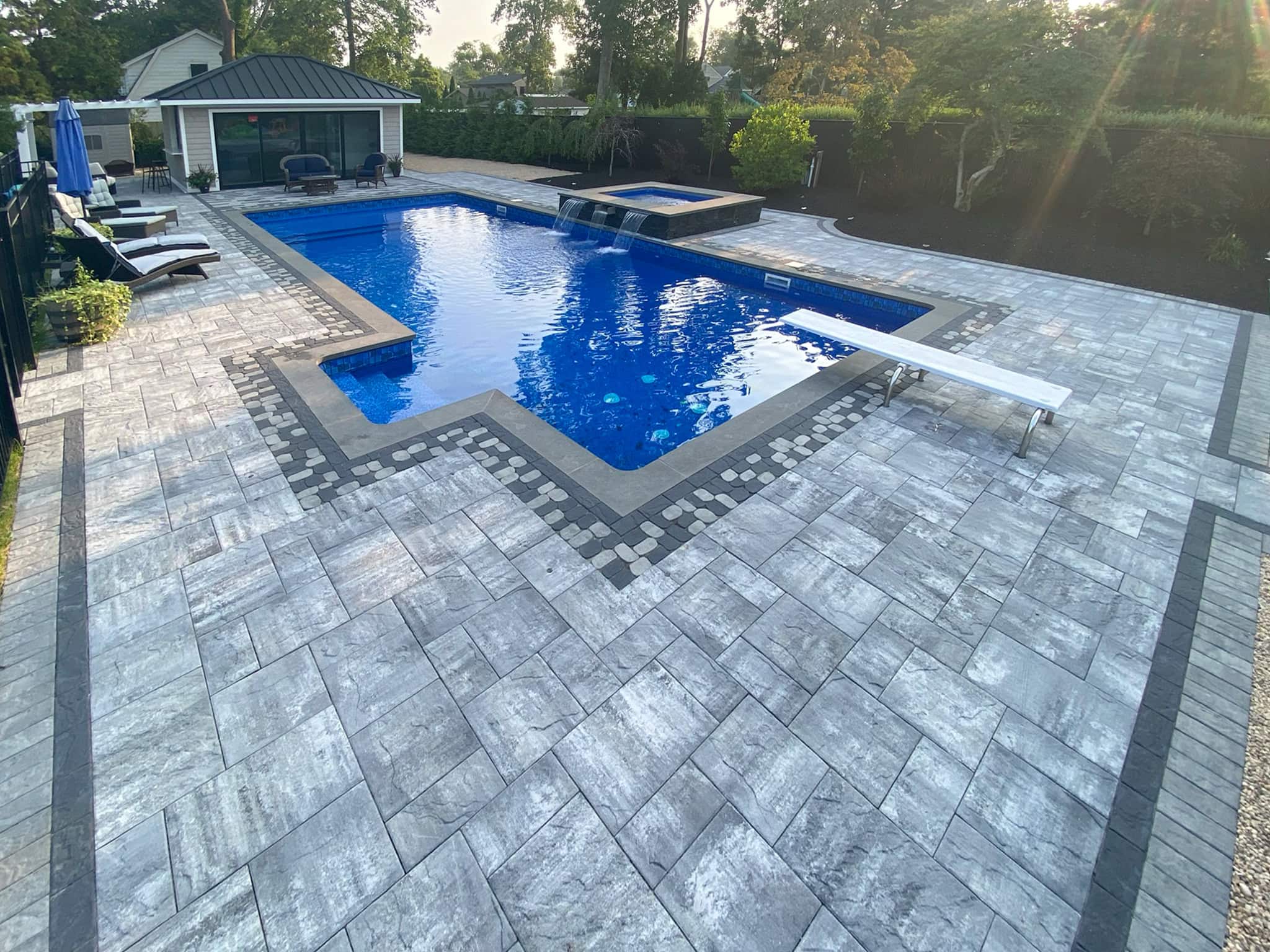 Rectangular swimming pool with a built-in hot tub, surrounded by gray stone pavers, lounge chairs, and a pool house in the background.