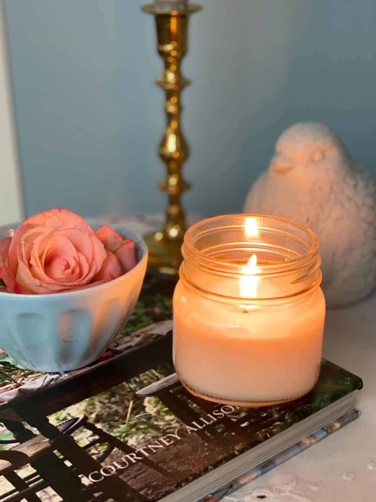 A lit candle in a glass jar sits on a book beside a small bowl with pink roses. A brass candle holder and a white bird ornament are in the background on a light-colored surface.