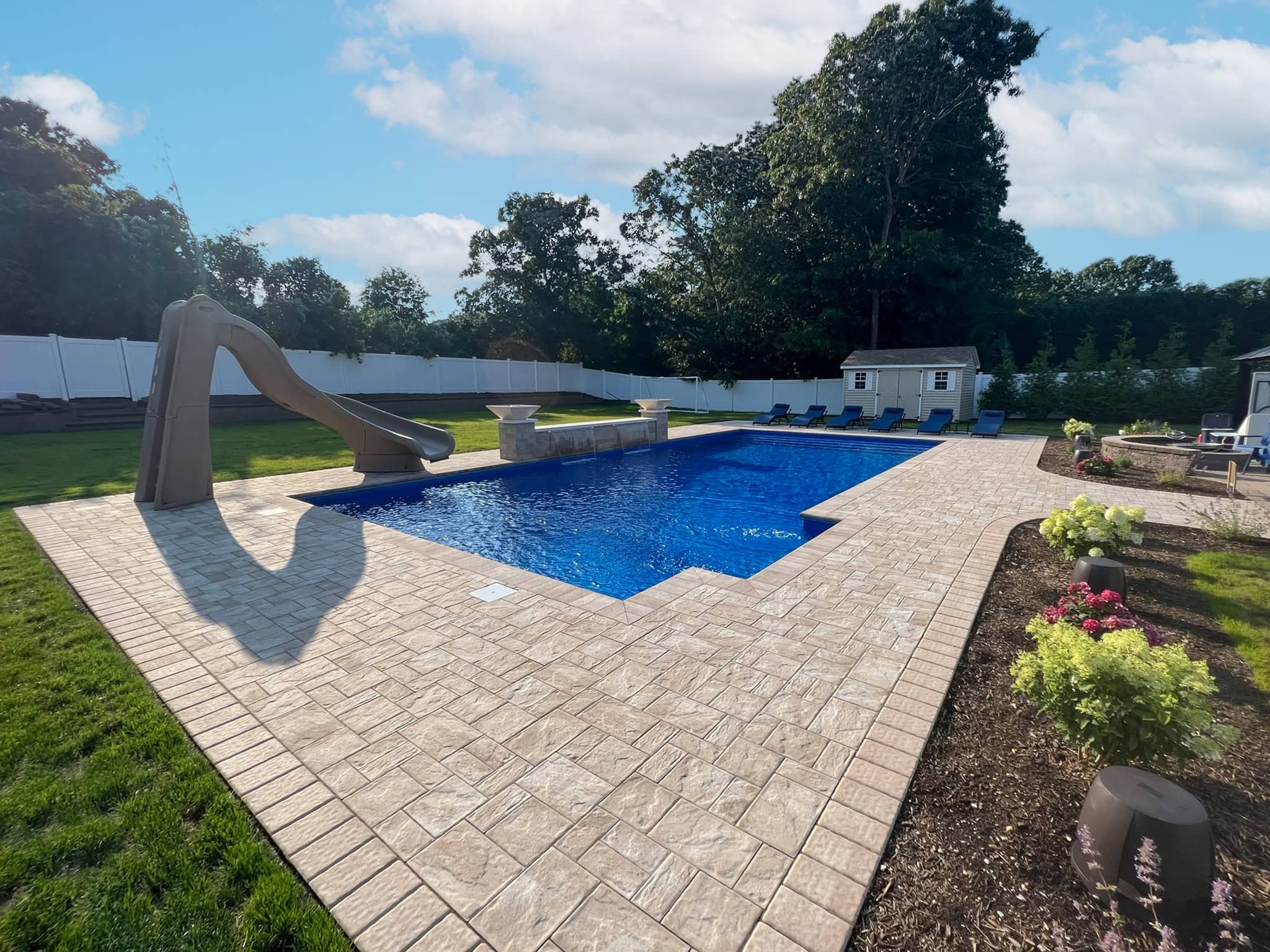 Rectangular backyard swimming pool with a slide, surrounded by a stone patio, lounge chairs, garden beds, and a white fence; trees and a shed in the background.