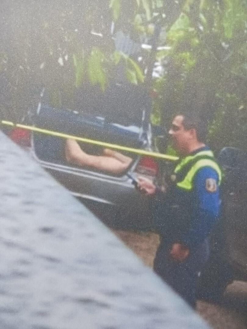 A crime scene in Costa Rica: A leg protrudes from the trunk of a car, cordoned off by yellow tape, with a police officer in uniform examining the scene. Possible evidence of Investor Fraud in a Real Estate Dispute.