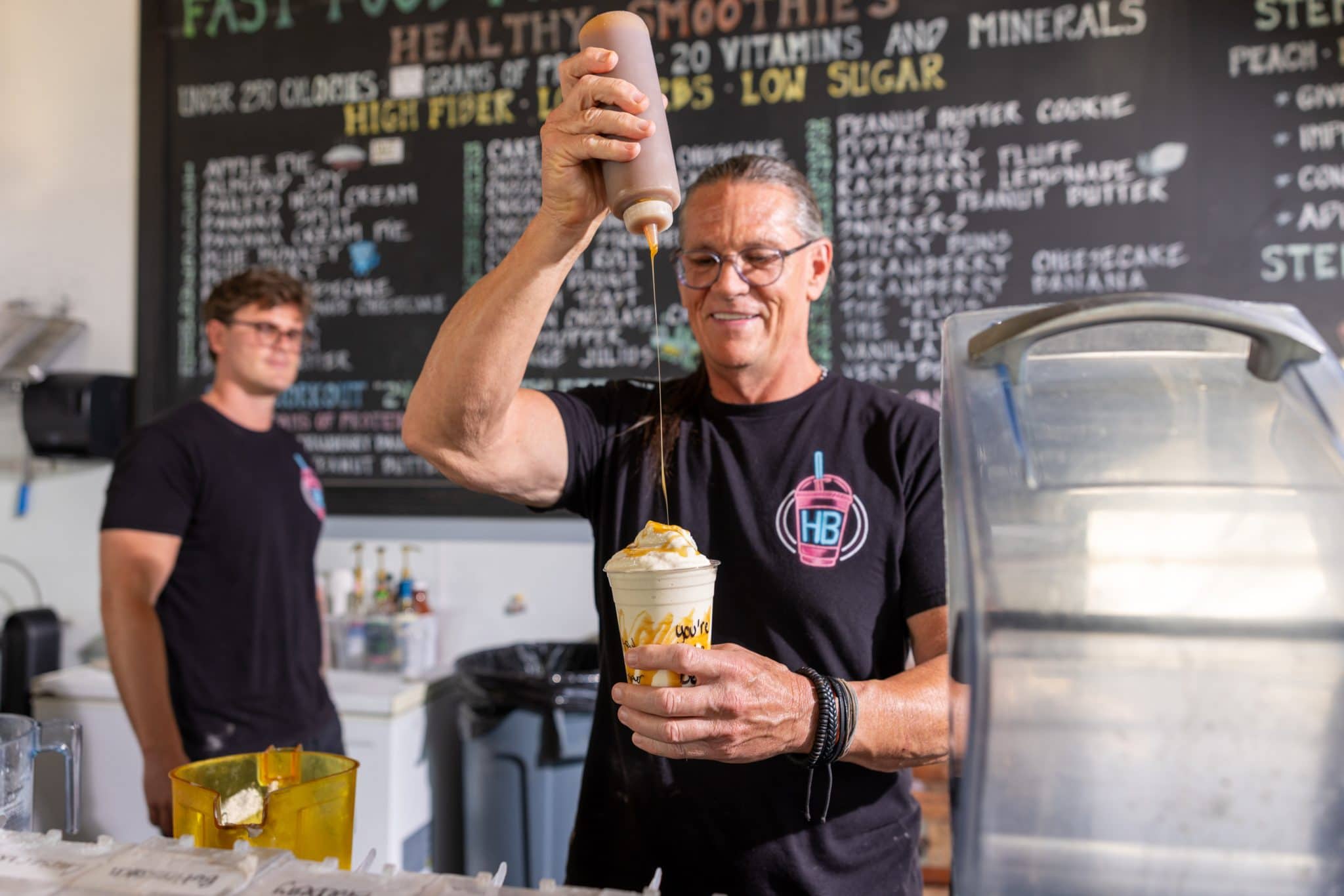 HB Protein Smoothies founder Daniel Boone finishing a shake at the Scottsdale location with the menu board visible behind him.