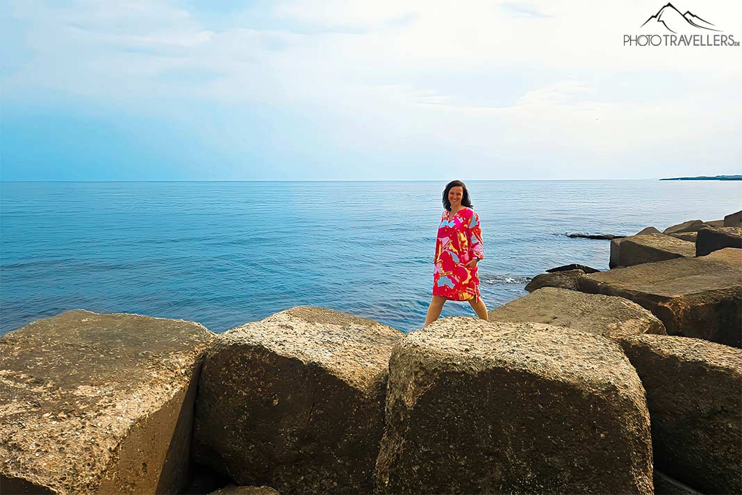 Eine Frau mit langen braunen Haaren in einem pinkfarbenen Kleid balanciert auf Steinquadern am Meer