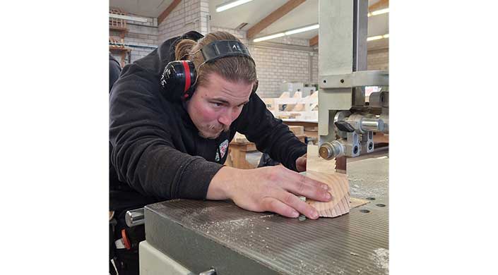 Ein Mann mit Kopfhörern arbeitet konzentriert an einem Holztisch in einer Werkstatt.