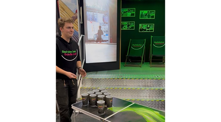 Mehrere Pappbecher stehen auf einem Tisch vor einer großen Leinwand, auf der eine Person im Hintergrund zu sehen ist.