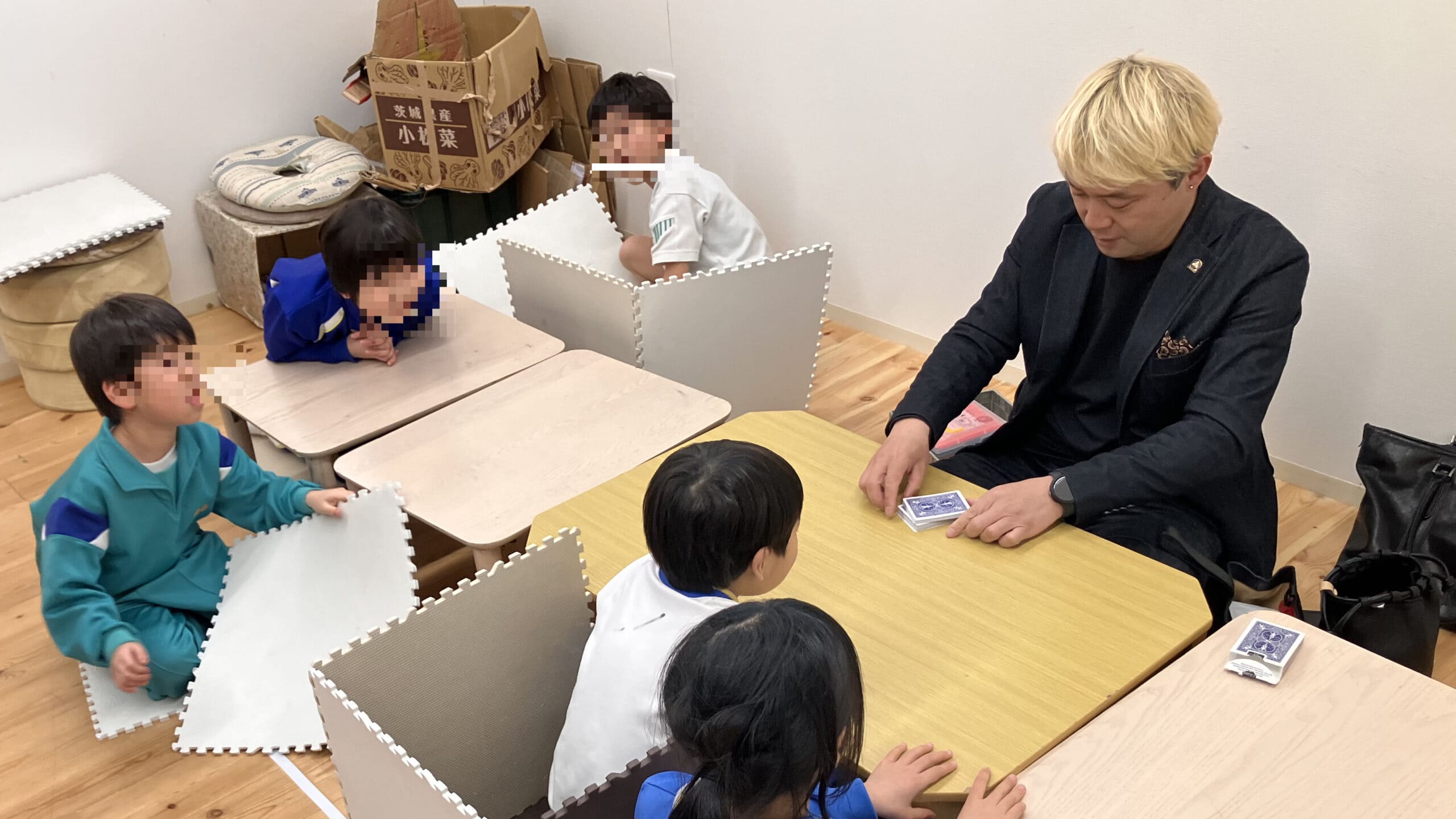 学童保育でマジックショー!折り紙マジックに40分かけて挑戦した子どもたち 1 学童保育の教室でテーブルを囲み、子どもたちにカードマジックを見せるマジシャン・コンプレッサー