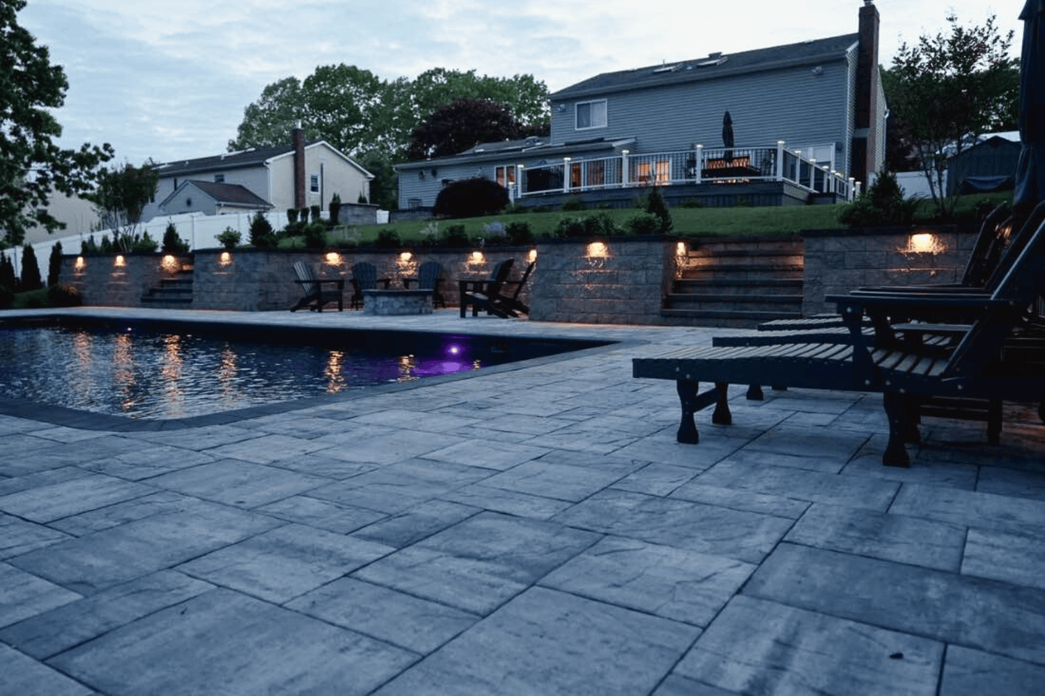 A backyard with a rectangular swimming pool, stone patio, lounge chairs, and tiered landscaping with retaining walls and lights, plus a house in the background.