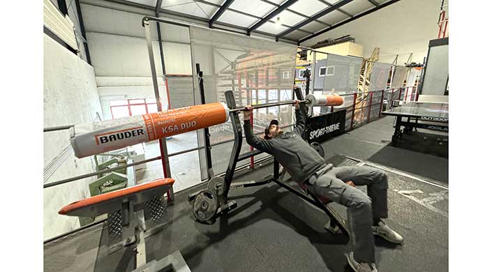 Eine Person liegt auf einer Bank und hebt eine Hantelstange mit einem orangefarbenen Schaumstoffpolster in einem Fitnessstudio.