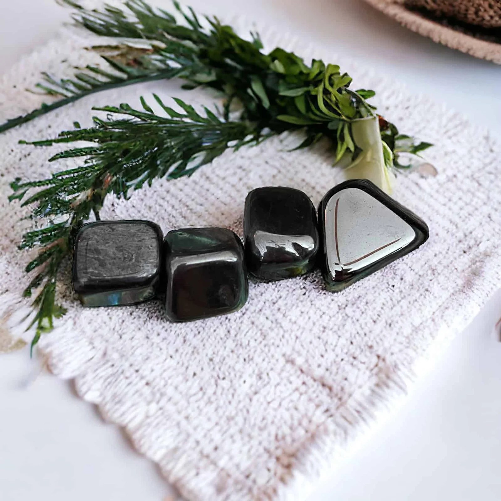 Four Hematite Tumble Stones are displayed on a textured white mat with green foliage, creating a calm and natural scene.