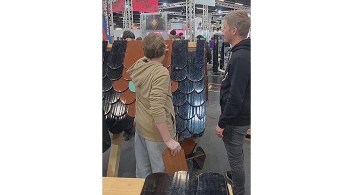 Ein Junge betrachtet bei einer Messe oder Ausstellung eine Wand mit Dachziegeln; weitere Menschen sind im Hintergrund sichtbar.