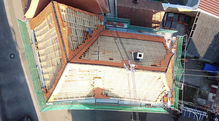 Bauarbeiter arbeiten auf einem Dach, das mit Holzbalken und Ziegeln gedeckt wird. Ein Kran hebt Baumaterialien an die Baustelle.