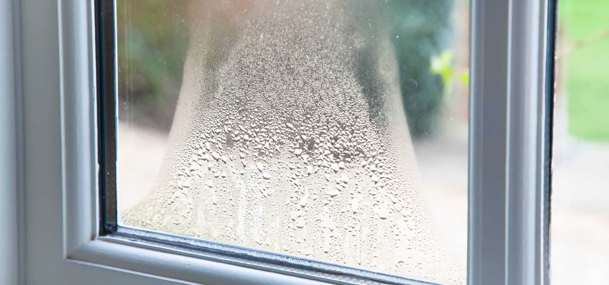 A close-up of a window pane with visible condensation, creating a misty effect that distorts the view outside. The window frame is white, and part of the green outdoors is faintly visible through the glass, almost as if a locksmith meticulously crafted an intricate blur across the scenery.