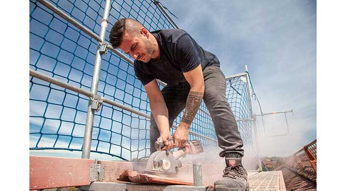 Ein Mann mit tätowiertem Arm benutzt eine elektrische Säge, während er sich über ein Metallgitter beugt, Staub steigt während der Arbeit auf.