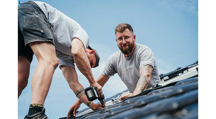 Zwei Männer installieren Solarpanels auf einem Dach, einer benutzt eine Bohrmaschine, beide tragen Arbeitskleidung, der Himmel ist klar und blau.