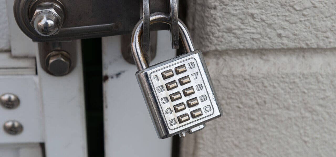 A closed combination padlock, securely fastened to a latch on a white, metal door, invites the skill of any seasoned locksmith. The lock's silver body is rectangular with a grid of numbered buttons for the combination. The surrounding wall appears textured and painted in a light color.