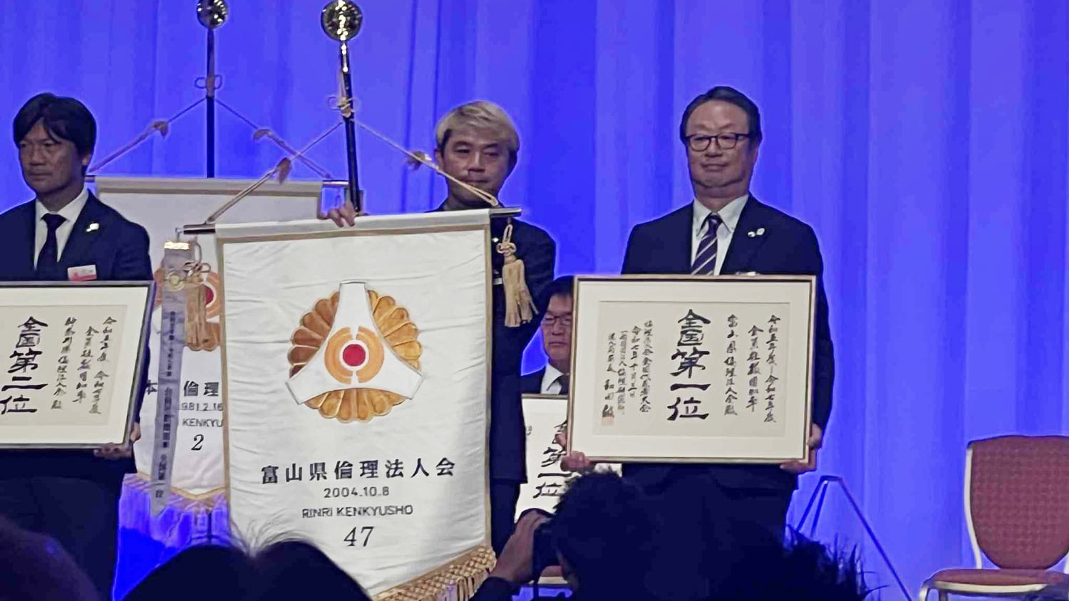 倫理法人会 全国代表者大会 富山県が全国一位の快挙!心でつながる経営者の学びの輪 6 富山県倫理法人会が全国第1位の表彰を受け、壇上で賞状と旗を掲げている様子