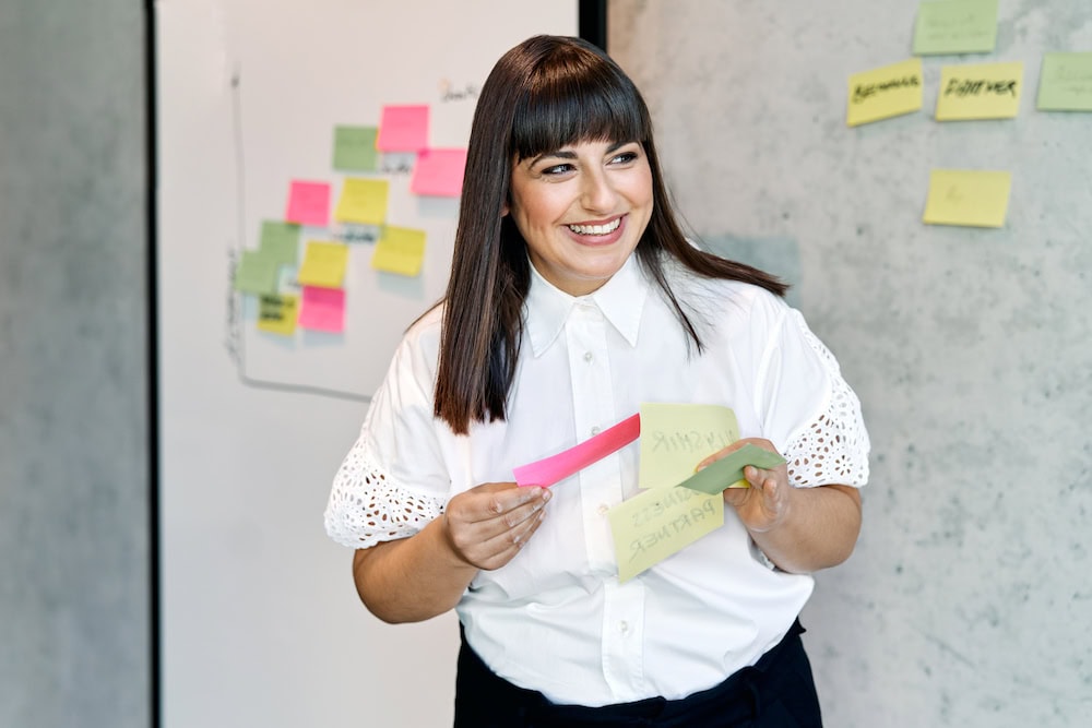 Floria Moghimi hält Post its in der Hand und steht vor einer Wand, auf der bereits Post its kleben.