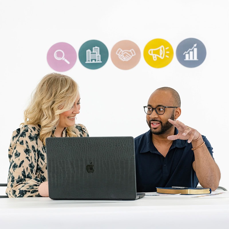 A marketing agency professional collaborates with a client at a laptop under strategy icons.
