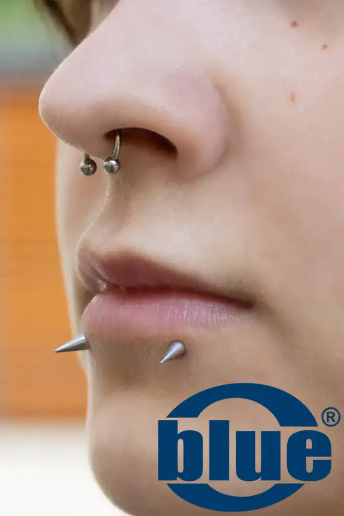 Close-up of a person’s side profile showing a silver nose hoop and lip piercings, with a blue logo partially overlaid at the bottom right.
