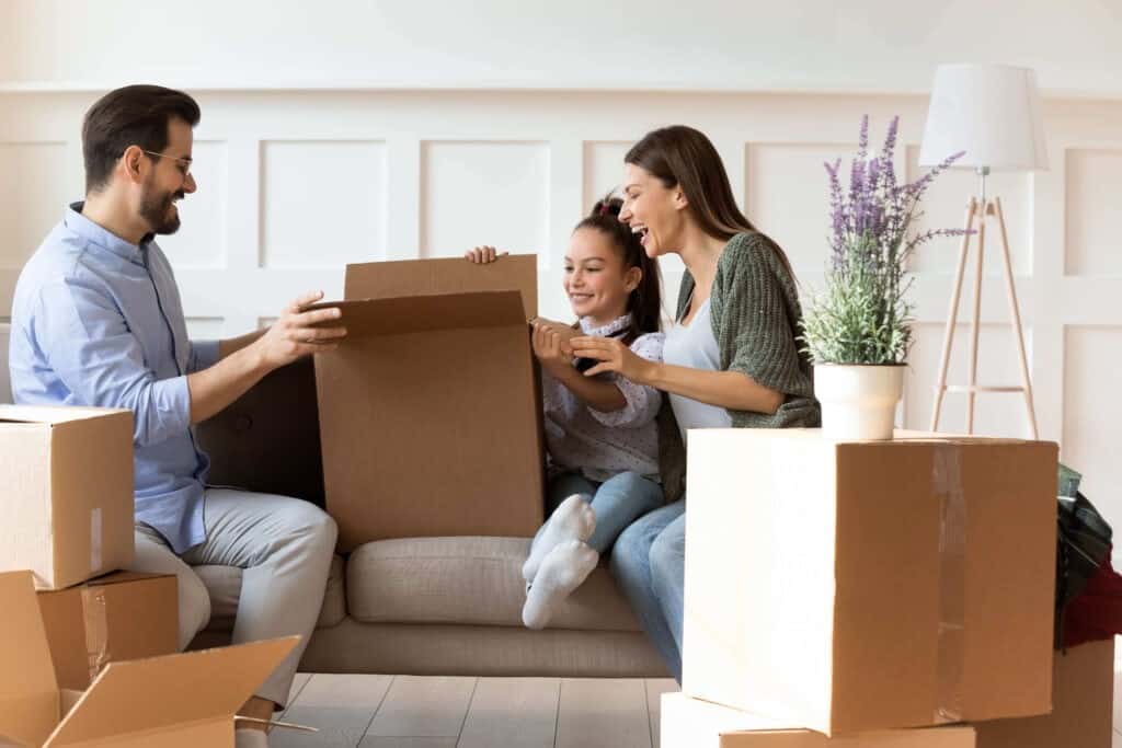 Adorable daughter helping parents with cardboard boxes on moving day, happy family with child sitting on couch, unpacking belongings in modern living room, relocation and mortgage concept