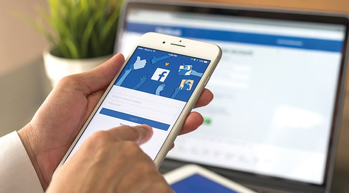 Eine Person hält ein Smartphone mit geöffneter Facebook-App, das Anmeldefenster und verschiedene Like-Symbole sind auf dem Bildschirm sichtbar.
