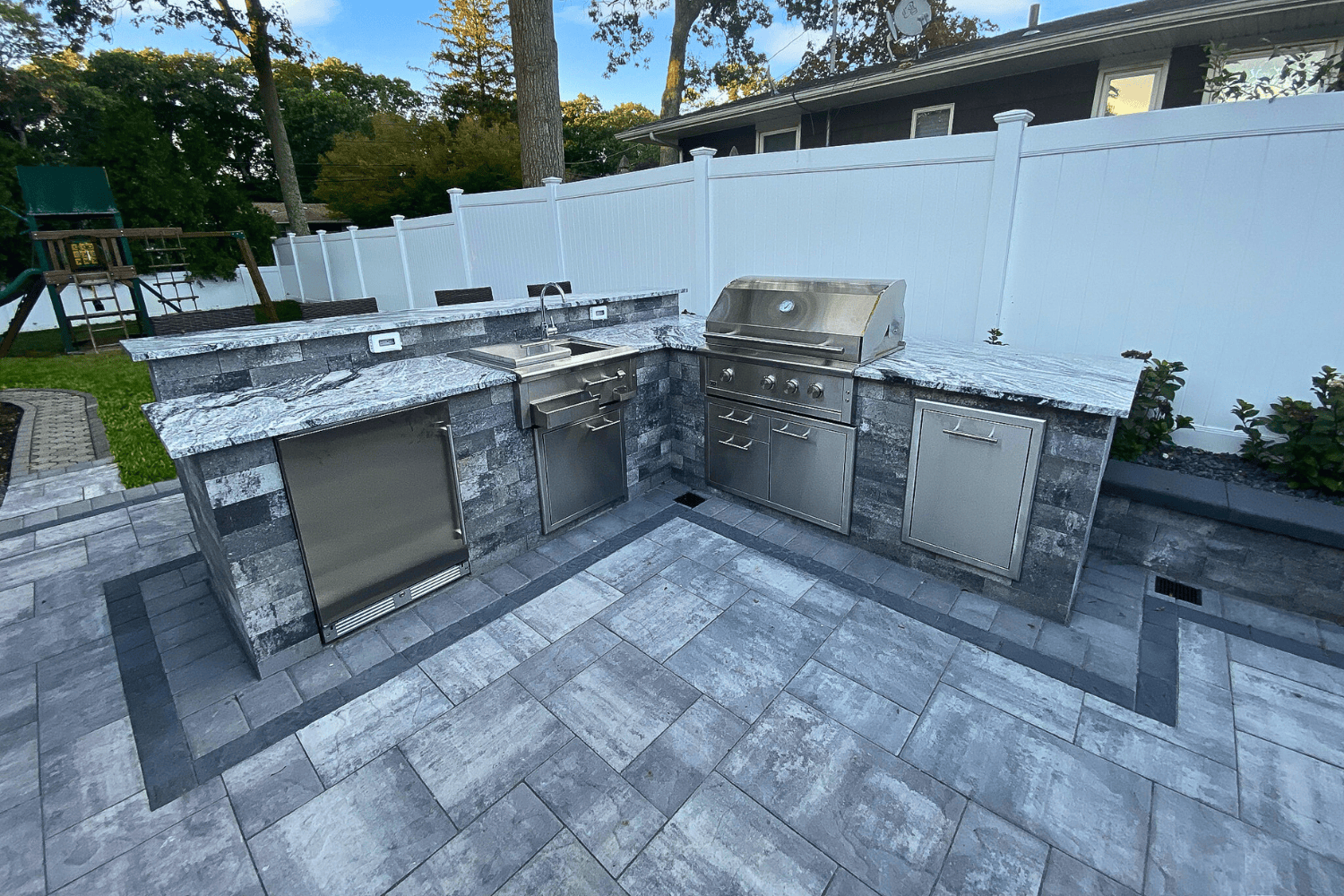 This Outdoor Kitchens setup features stainless steel appliances—grill, sink, and fridge—in a stone countertop and cabinetry, all on a paved patio with a white fence in the background.