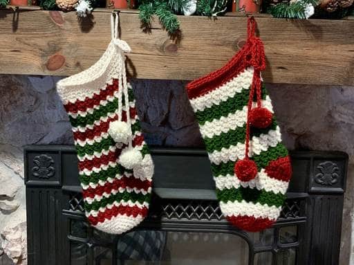 Two Christmas stockings with red, green, and white stripes and two pom-poms each hang from a wooden mantel above a fireplace. The stockings have a crocheted texture; no visible text or promotional info appears in the image. Free crochet pattern