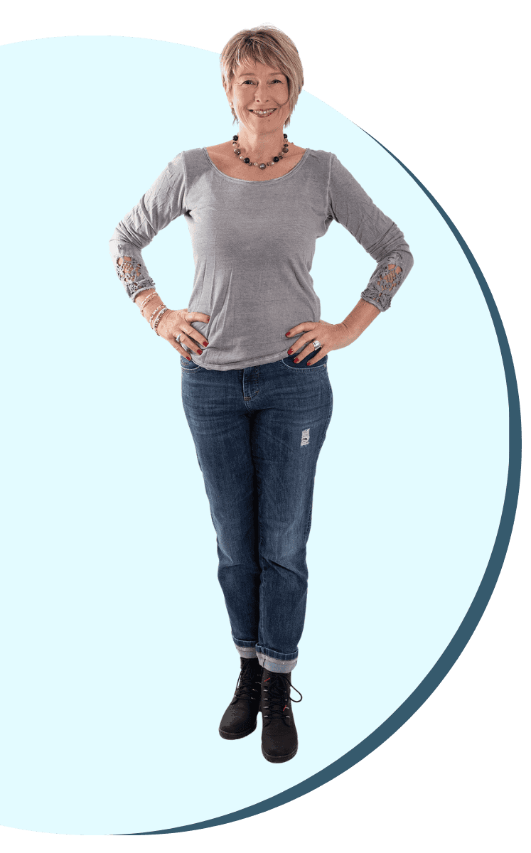 Sabine Piarry in grauem Langarmshirt und Jeans steht mit in die Hüfte gestützten Armen vor einem blau-weißen grafischen Hintergrund.