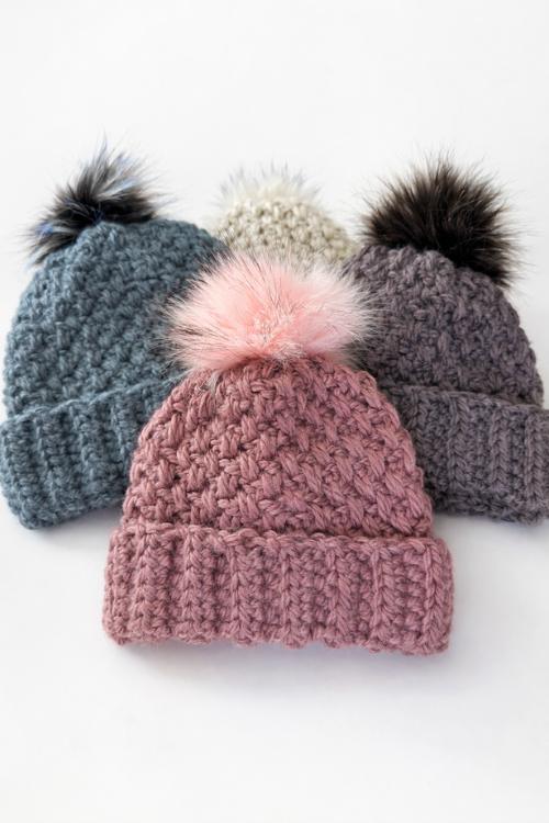 Four textured beanies with fluffy pom-poms in blue, white, gray, and pink are arranged on a white surface. No text visible. Free crochet pattern