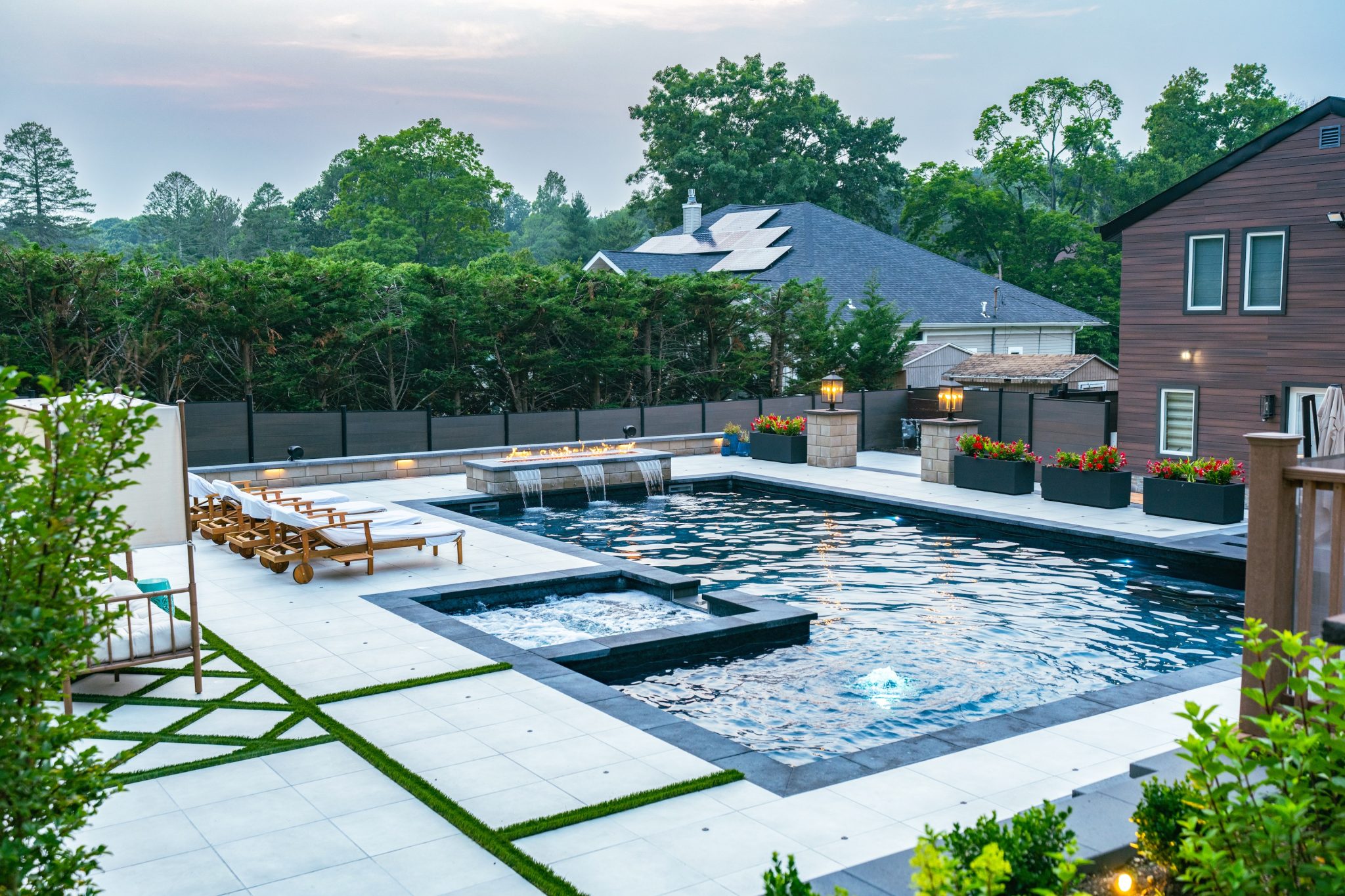 Modern backyard with a large rectangular swimming pool, lounge chairs, spa area, landscaped greenery, and surrounding privacy fence.