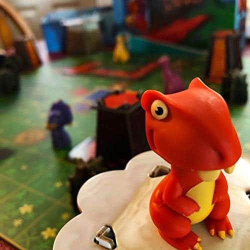 Petite figurine de dinosaure rouge en plastiques dans un jeu de société ou décor, avec un fond coloré et des éléments de jouets et d’activités pour enfants.