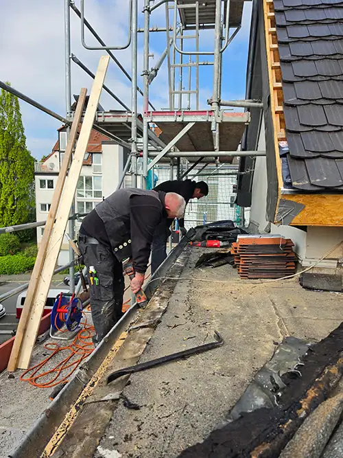 Zwei Dachdecker arbeiten auf einem Gerüst an einem Dach, umgeben von Werkzeugen und Baumaterialien, während sie Sanierungsarbeiten durchführen.