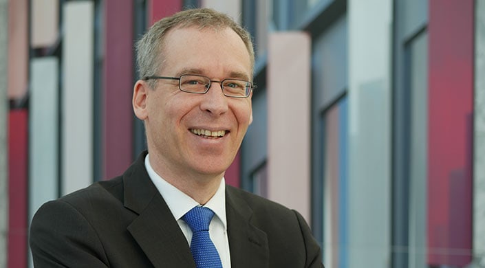 Ein Mann im Anzug und mit Brille lächelt in die Kamera, Hintergrund unscharf mit modernen Gebäuden.